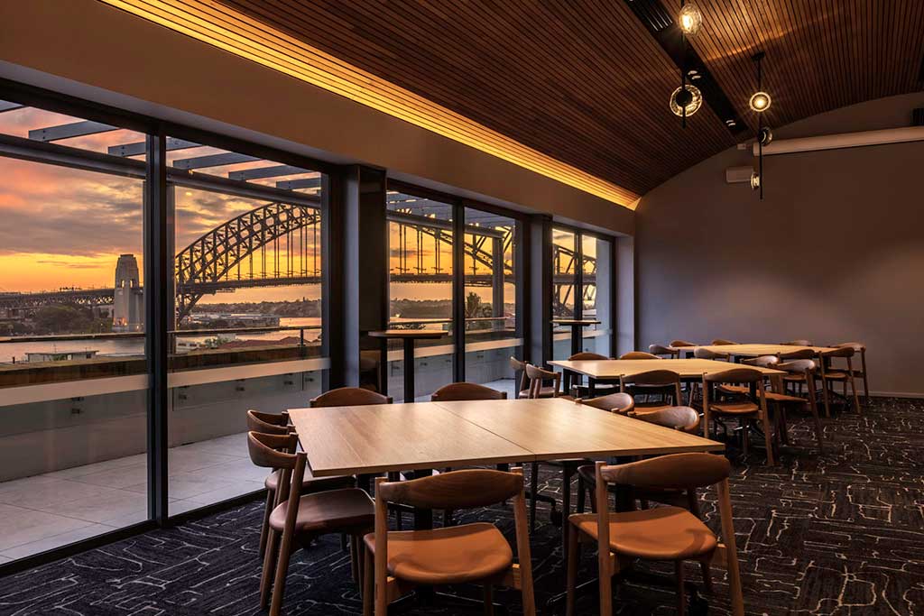 Level 4 dining room with a large balcony overlooking a view of the Sydney Harbour Bridge at dusk. Square dining tables with 8 chairs are laid out in the room.